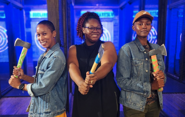 three smiling women holding axes at an axe throwing club.